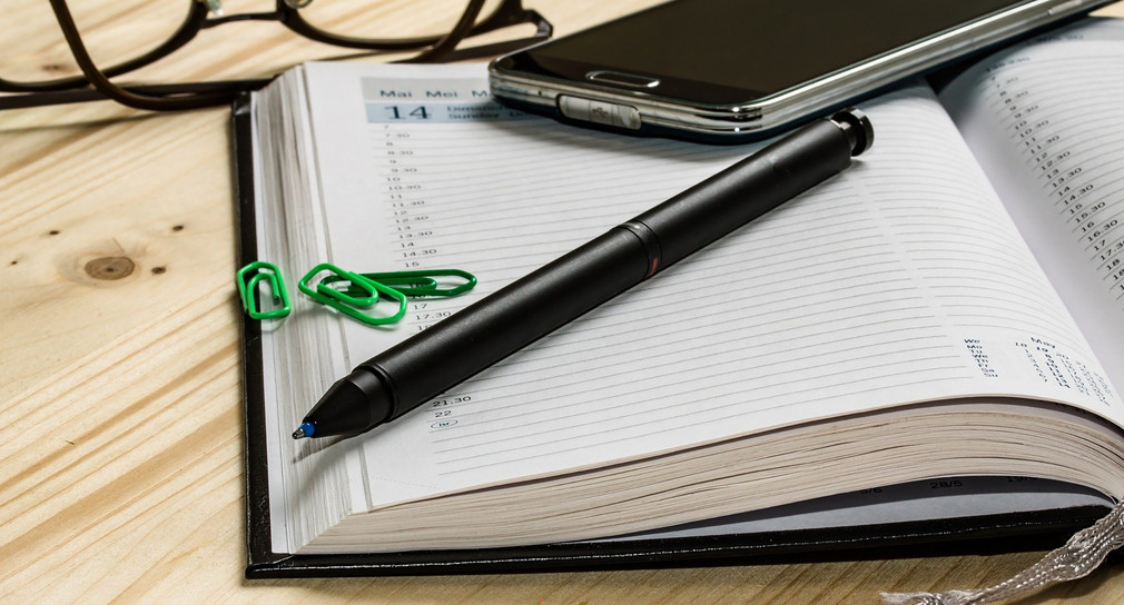 Ein Terminkalender, ein Handy, eine Brille und ein Stift liegen auf dem Tisch