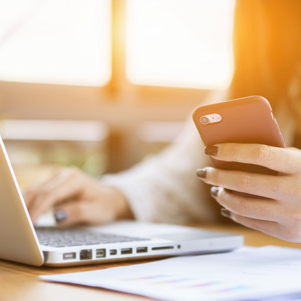 Symbolbild: Frau am Laptop mit Smartphone in der Hand