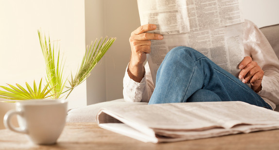 Beschreibung vom Bild: Man sieht einen Mann. Er sitzt auf der Couch. Er liest eine Zeitung. 