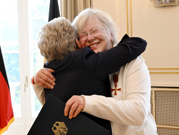 Auszeichnung Bundesverdienstkreuz für Edeltraut Stiedl Zwei ältere Frauen umarmen sich herzlich, eine hält eine Mappe mit dem deutschen Bundesadler, im Hintergrund eine deutsche Flagge.