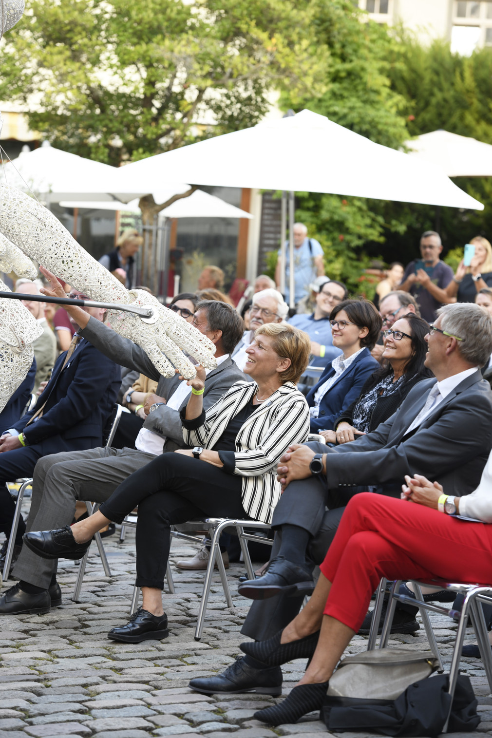 Ministerin Nicole Razavi MdL sitzt gemeinsam mit weiteren Gästen auf dem Platz vor dem Schloss in Meersburg und eine übergroße Figur - DUNDU - reicht Ihr die Hand
