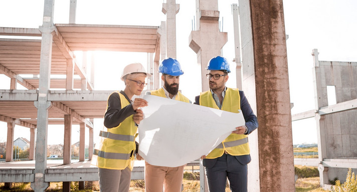 Eine Prüfingenieurin und zwei Prüfingenieure begutachten einen Plan auf einer Baustelle