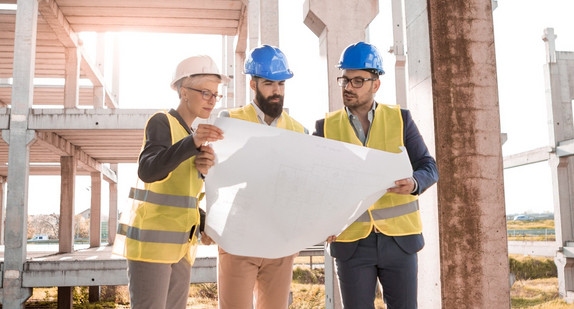 Eine Prüfingenieurin und zwei Prüfingenieure begutachten einen Plan auf einer Baustelle