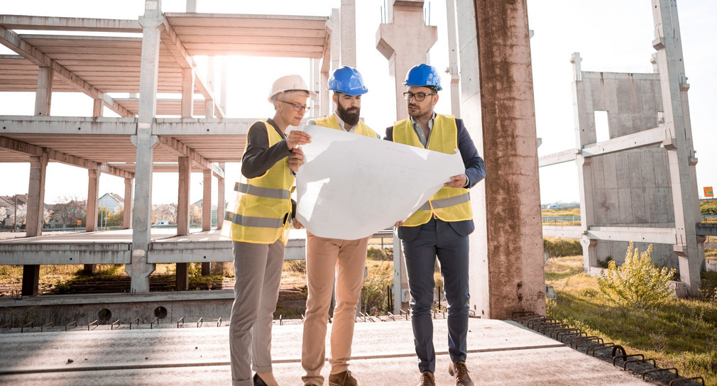 Eine Prüfingenieurin und zwei Prüfingenieure begutachten einen Plan auf einer Baustelle