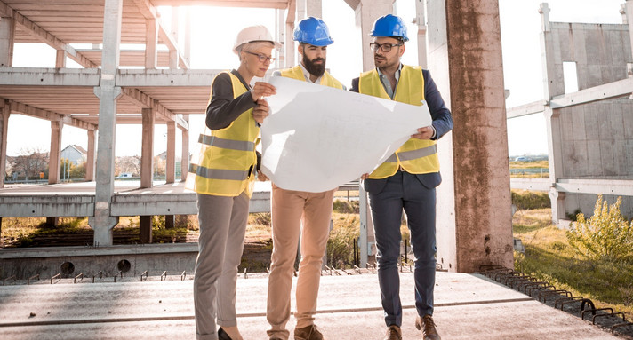 Bautechnik und Bauökologie Anerkennung als Prüfingenieurin und Prüfingenieur für Bautechnik Eine Prüfingenieurin und zwei Prüfingenieure begutachten einen Plan auf einer Baustelle
